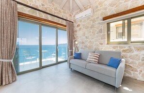 Ein Zimmer mit einer Steinwand, einem grauen Sofa mit Kissen und einer großen Schiebeglasdoor, mit Blick aufs Meer.