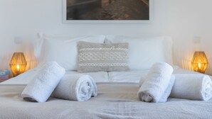 Un lit bien fait, avec des serviettes blanches roulées, un oreiller blanc et un tableau représentant un coucher de soleil sur une plage.