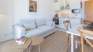 Un salon moderne comprenant un canapé, une petite table ronde avec des livres, une table à manger avec des chaises, une cuisine avec des meubles blancs et un luminaire suspendu.