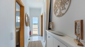Un couloir avec une porte en bois donnant sur un balcon, une commode blanche avec un miroir et une décoration en forme d’arbre fixée au mur.