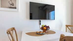 Un coin repas avec une table blanche, des chaises, un saladier de fruits et un tableau encadré au mur.