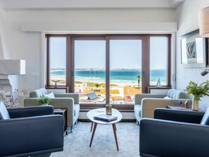 Ein modernes Wohnzimmer mit großem Fenster mit Blick auf einen Strand und einen Steg. Es gibt zwei Sofas, einen Couchtisch und eine Lampe.