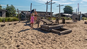 A sandy beach area with wooden play structures and a building in the background. - Duinlodge 4 + 2 (Noordwijk)