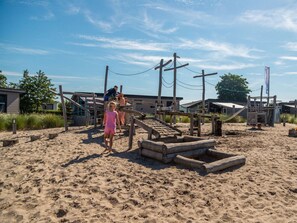 A sandy beach area with wooden play structures and a building in the background. - Duinlodge 4 + 2 (Noordwijk)