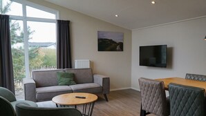 A modern living room with a sofa, a round coffee table, and a dining table with chairs. A TV is mounted on the wall. - Duinlodge 4 + 2 (Noordwijk)