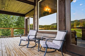 Eine Holzterrasse mit zwei Schaukelstühlen und einem Tisch, mit Blick auf einen See und Hügel.