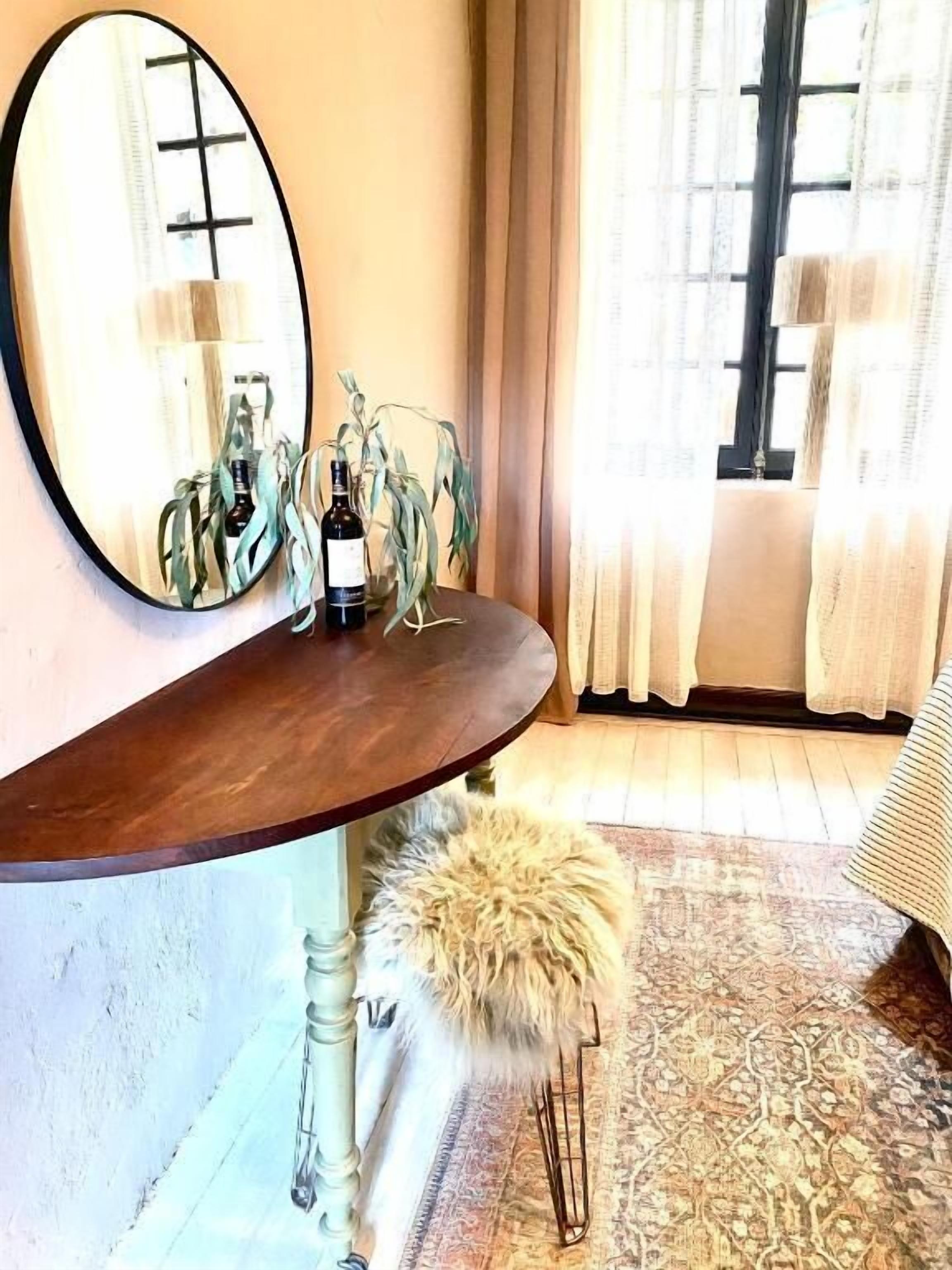 A room with a round mirror, a small table with bottles, a fluffy chair, and a patterned rug.
