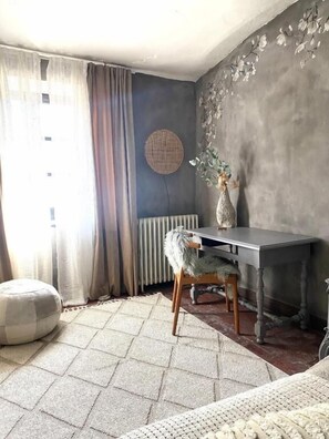 A room with a dark wall, a round woven wall hanging, a table with a chair, a radiator, and a window with curtains.