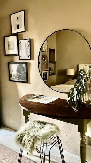 A room with a round mirror, a wooden table, a chair with a fur cushion, and framed pictures on the wall.