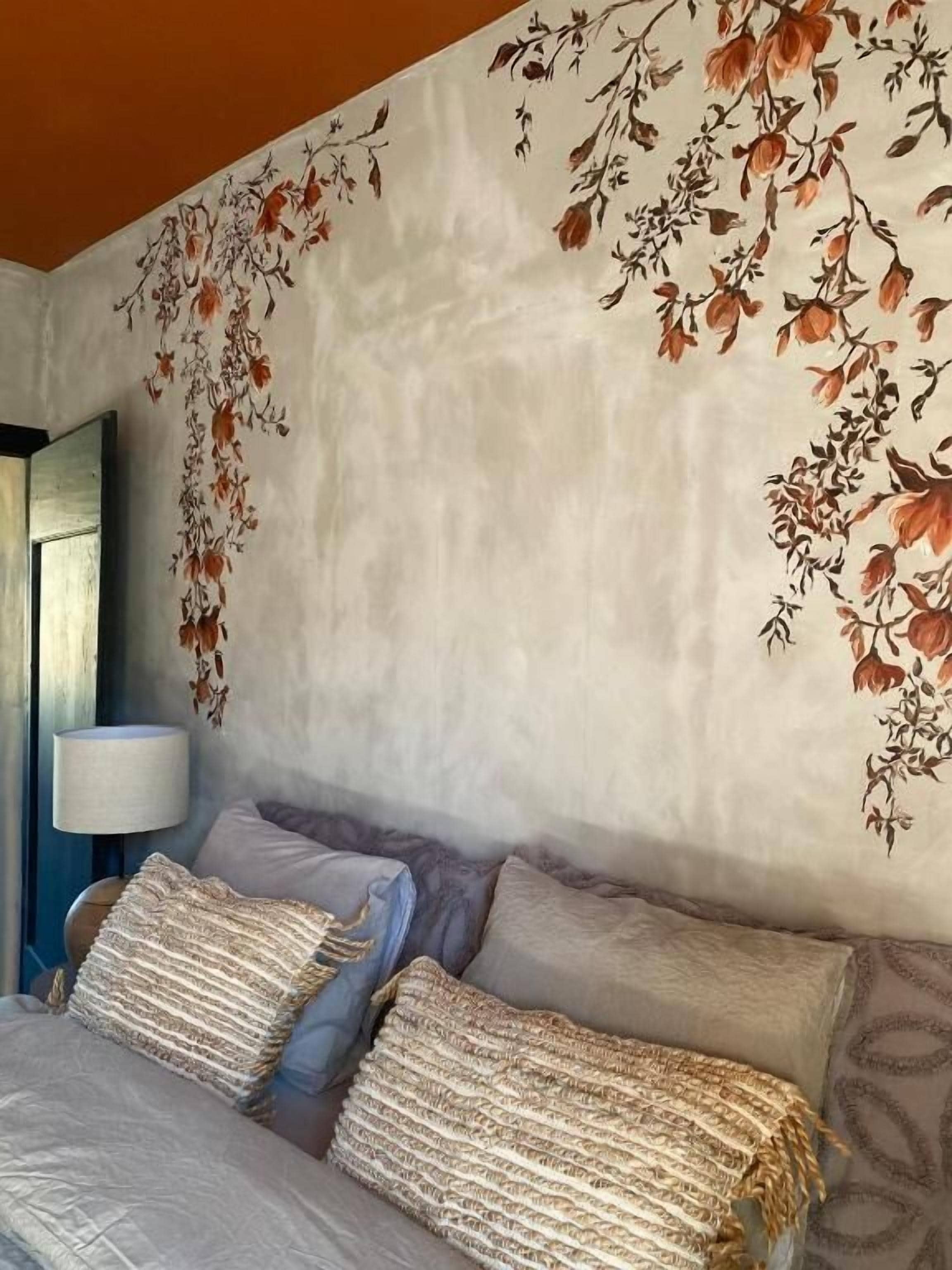 Un dormitorio con cama, almohadas, una lámpara y un mural floral en la pared.