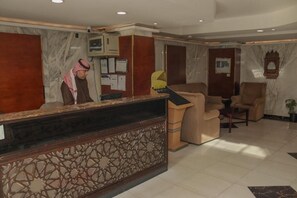 A hotel reception area with a marble reception desk, a man standing behind it, and several beige armchairs. - Al Orouba Hotel (Riyadh)