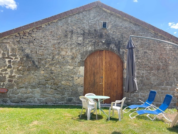 A stone building with a wooden door, outdoor seating with chairs and a table, and a lamp.