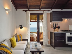 Ein gemütliches Zimmer mit einer Couch, einem Tisch, einer Küche mit Herd und Blick aufs Meer.