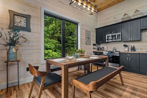 A wooden dining table with chairs, a kitchen with dark cabinets, a clock, and a large window with a view of greenery. - Mountain View Lodge, Hot Tub, Fire Table & Theater (Pigeon Forge)