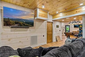 A cozy living room with a large flat-screen TV mounted on the wall, a wooden ceiling, and a comfortable sofa. - Nice Views, Theater, Gameroom, Arcade, Hot Tub (Pigeon Forge)