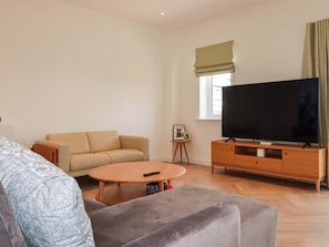 A living room with a sofa, a coffee table, a TV on a wooden stand, and a small side table with a picture frame. - Eden's Bloomdale (St Austell)