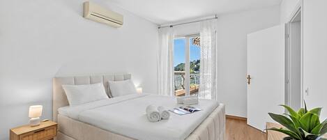 A bedroom with a bed, bedside table, lamp, curtains, and a potted plant.