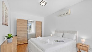 A modern bedroom with a large bed, a bedside table with a lamp, a potted plant, and a view through a sliding door. - Lugano Sun & View Apartment for 4 (Lugano)