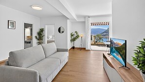 A modern living room with a grey sofa, wooden flooring, a flat-screen TV, and a view of a mountain landscape through a large window. - Lugano Sun & View Apartment for 4 (Lugano)