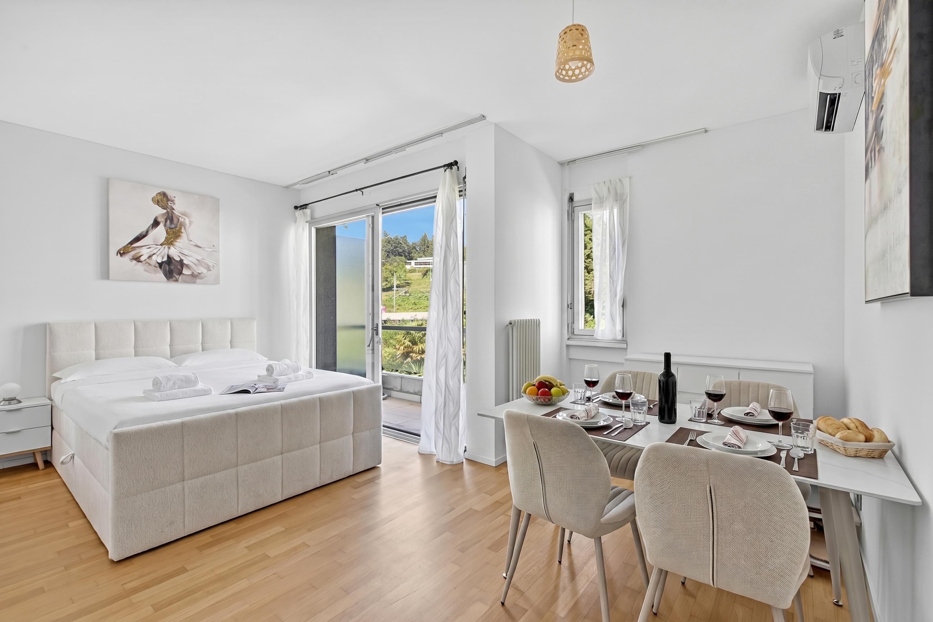 A modern bedroom with a large bed, a dining area with a table set for two, and a sliding glass door leading to a balcony.