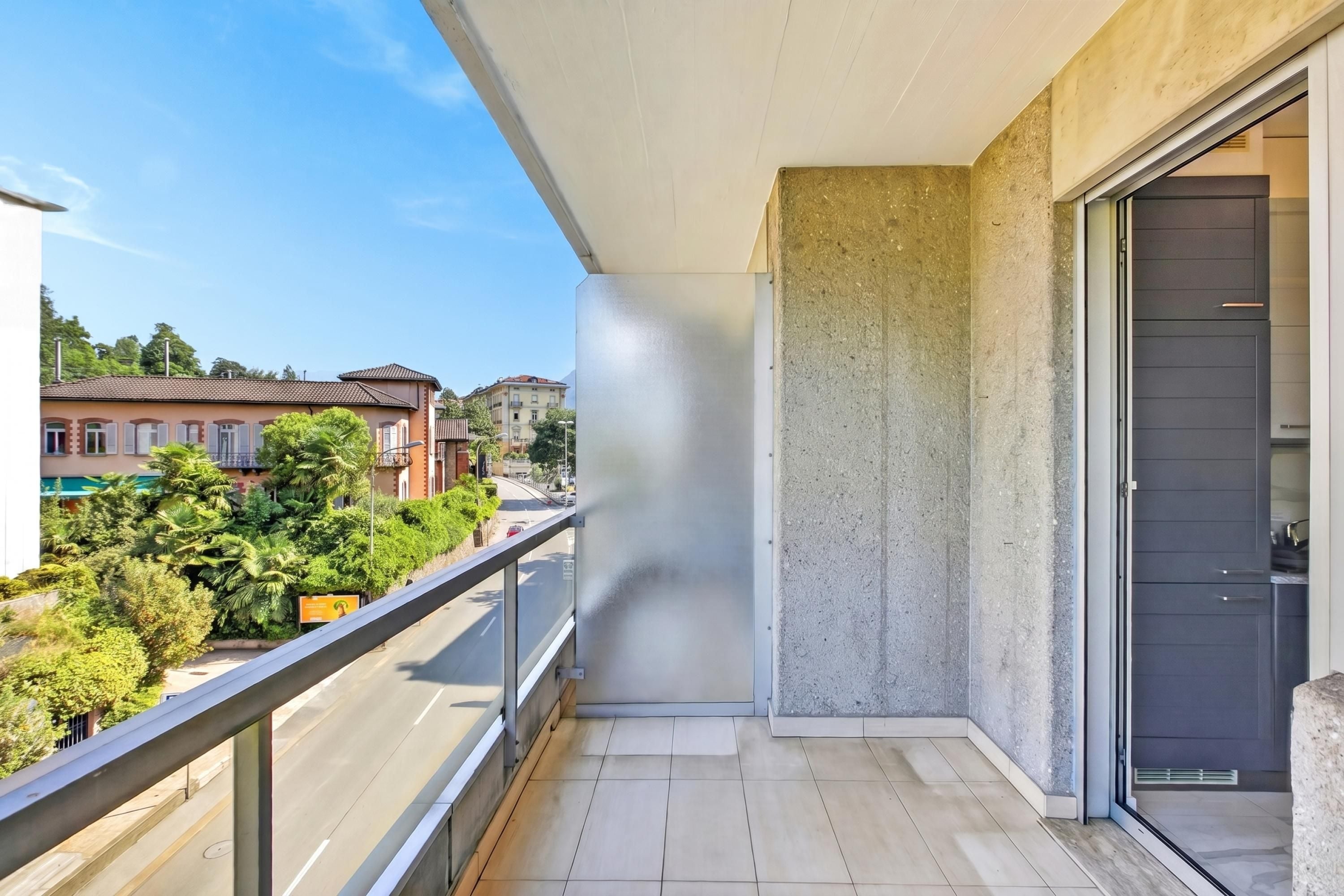 Een balkon met uitzicht op een straat, een betonnen muur en een schuifdeur van glas.