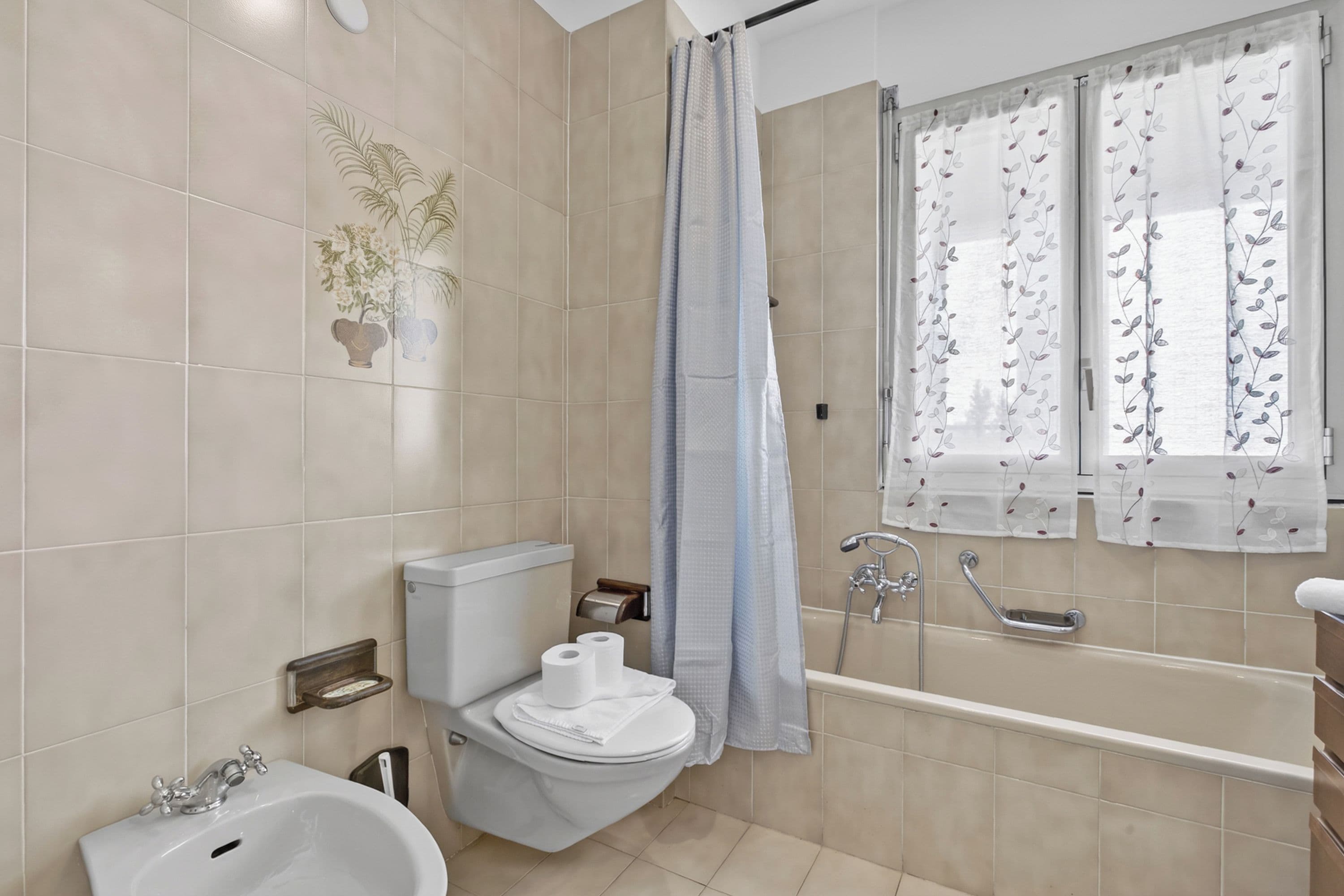 Un baño con inodoro, bidé, bañera y ventana con cortina floral.