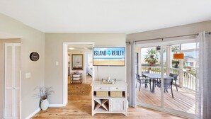 Een interieur met een televisie waarop 'ISLAND REALTY' staat, een houten vloer, een eetgedeelte met buitenterras en uitzicht op het strand.