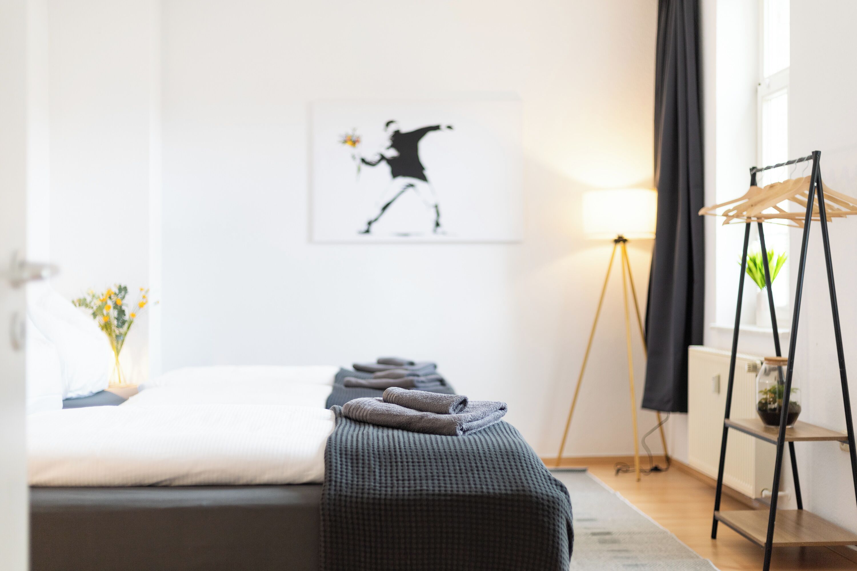 Ein ordentlich eingerichtetes Schlafzimmer mit einem Bett, einem Nachttisch mit Lampe, einem Wandbild und einem Kleiderständer.