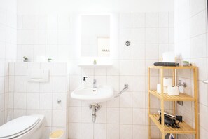 Kylpyhuone, jossa on wc, pesuallas, peili ja hyllyjä, joilla on hygieniatuotteita.