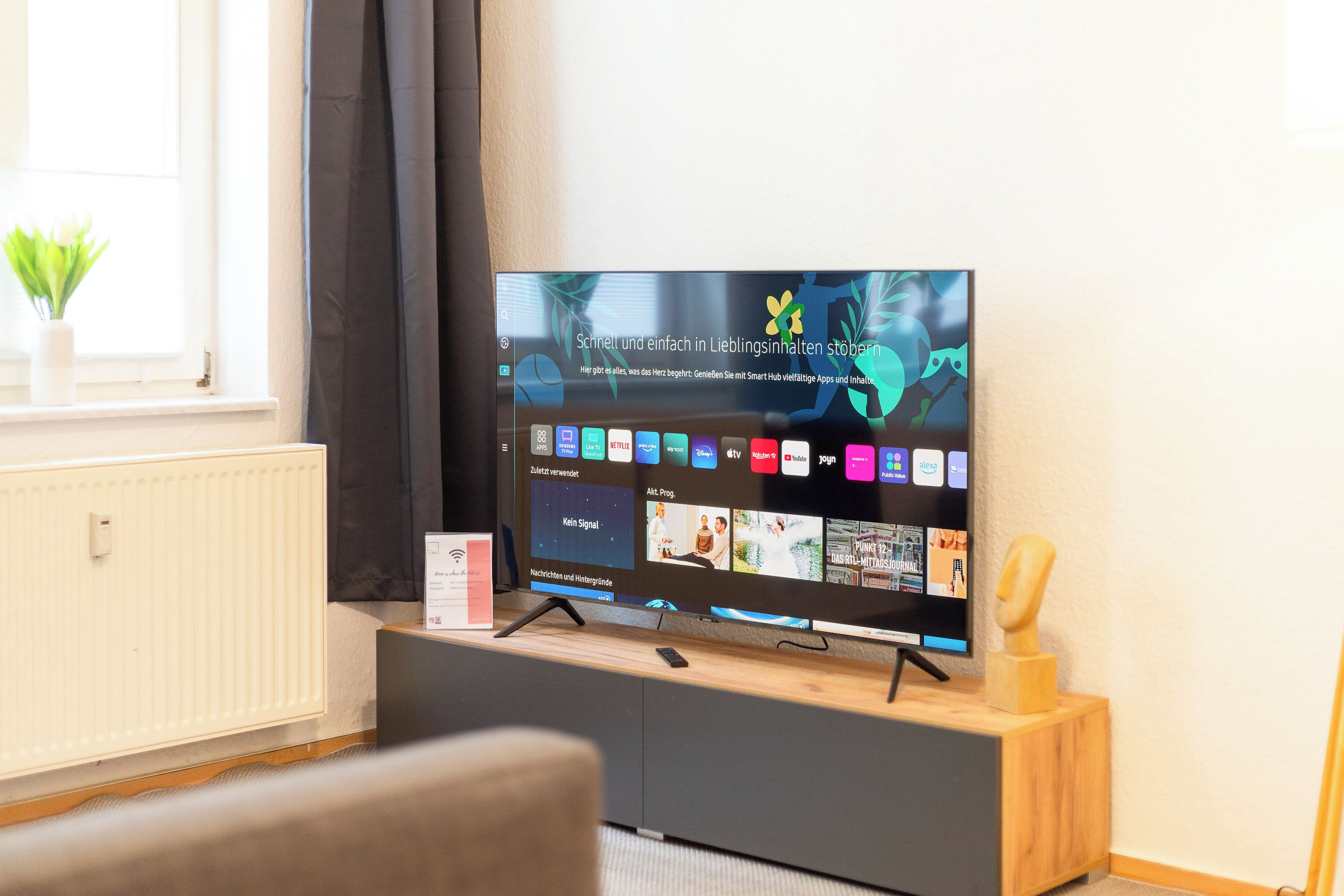 A flat-screen TV on a wooden stand in a room with a window, a radiator, and a small plant.
