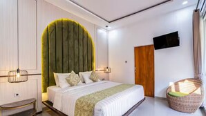 A modern bedroom with a curved headboard, a bed with white bedding, a wicker chair, and a flat-screen TV on the wall. - 2BR Villa w private Pool - Queen (Seminyak)