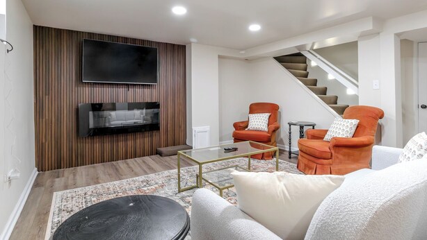 Una sala de estar moderna con un televisor de pantalla plana, dos sillones naranja, una mesa de centro de vidrio y una escalera.