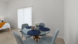 A modern room with a glass dining table, grey chairs, a bed with an orange throw, and a white wall. - Garage Studio King + Queen Studio | Private Patio (Smithfield)