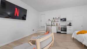 A modern, compact living space with a kitchenette, a TV mounted on the wall, a bed with an orange throw, and a seating area with a TV. - Garage Studio King + Queen Studio | Private Patio (Smithfield)