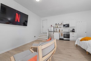 Ein modernes, kompakter Wohnraum mit Küchenzeile, einem an der Wand befestigten Fernseher, einem Bett mit orangefarbener Decke und einer Sitzecke mit Fernseher.