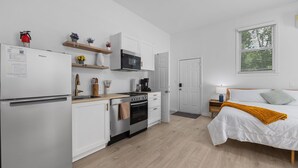 A compact kitchen with white cabinetry, a microwave, and a stove. There is a bed with a white comforter and a mustard throw blanket. A small nightstand with a lamp is next to the bed. - Garage Studio King + Queen Studio | Private Patio (Smithfield)