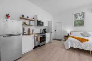 Eine kompakte Küche mit weißen Schränken, einer Mikrowelle und einem Herd. Ein Bett mit weißer Bettdecke und einem senffarbenen Überwurf. Ein kleiner Nachttisch mit Lampe steht neben dem Bett.