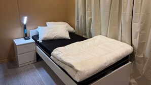 Un lit simple avec un matelas noir et du linge de lit blanc, une table de chevet avec une lampe et un rideau qui recouvre une fenêtre.