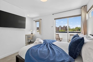Ein Hotelzimmer mit Bett, Schreibtisch, Stuhl, Fernseher und einem Balkon mit Blick auf Gebäude.