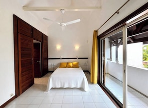 Ein Schlafzimmer mit Bett, Deckenventilator, einer Schiebetür aus Glas und Blick auf Grünflächen.