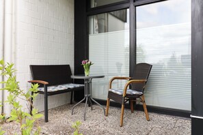 Een terras met een zwart metalen bankje, een rieten stoel en een kleine tafel met een bloevas.