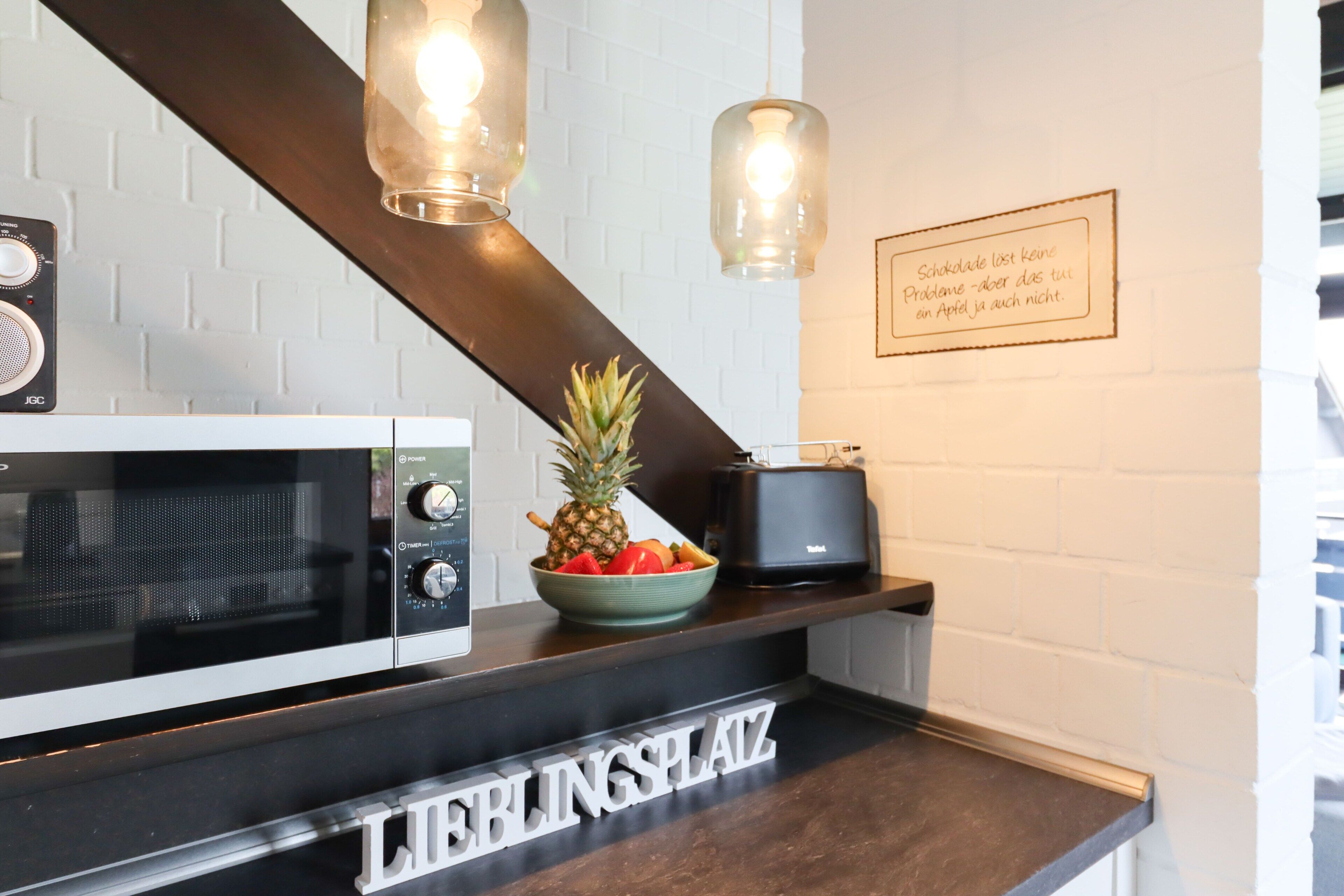Un plan de travail de cuisine avec un micro-ondes, un saladier de fruits, un grille-pain et une pancarte sur laquelle on peut lire « LIBLINGSPLAZ ».