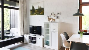 Una sala moderna con televisor de pantalla plana, un mueble blanco con puertas de cristal, una mesa de comedor con sillas y una ventana con cortinas.