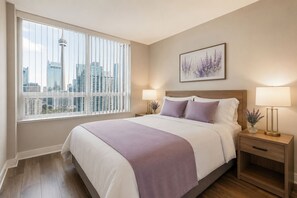 Ein Schlafzimmer mit einem Bett, zwei Nachttischen mit Lampen, einem Fenster mit Jalousien, Blick auf die Stadt und einem gerahmten Kunstwerk an der Wand.