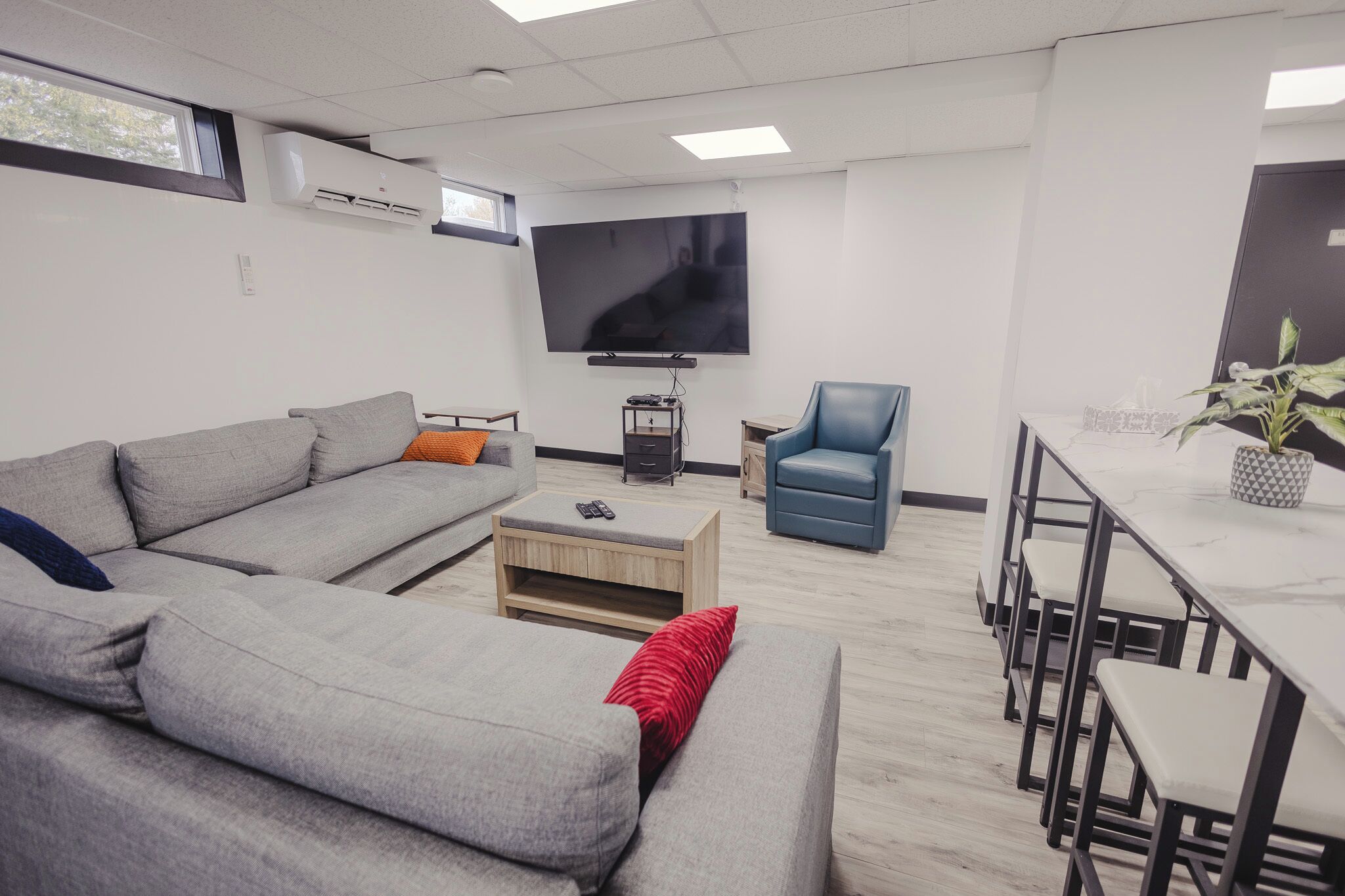 Una sala de estar moderna con un sofá gris, un sillón azul, un televisor y un comedor con mesa y sillas.
