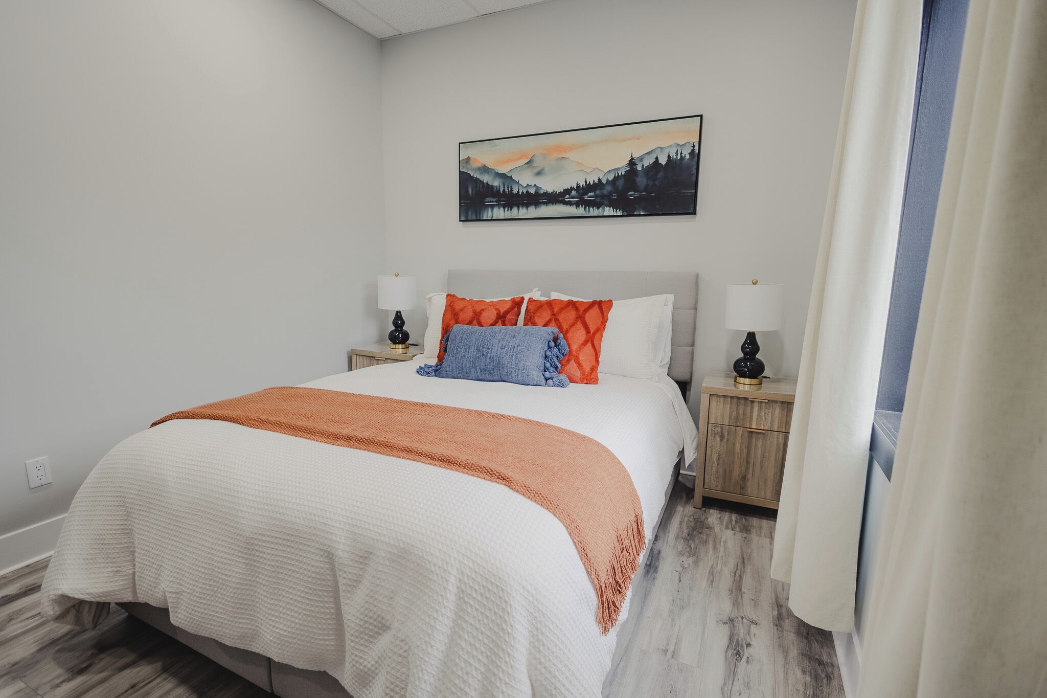 A neatly made bed with a white and orange blanket, two bedside lamps, a nightstand, and a painting of a mountain landscape on the wall.
