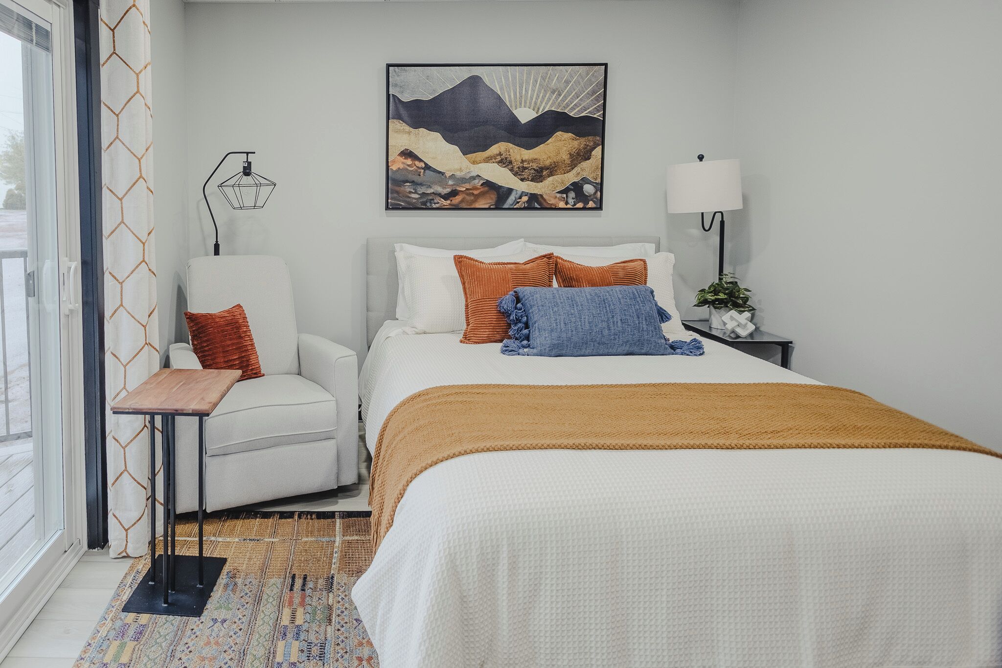 A bedroom with a bed, a chair, a nightstand, a lamp, a painting, and a window with curtains.