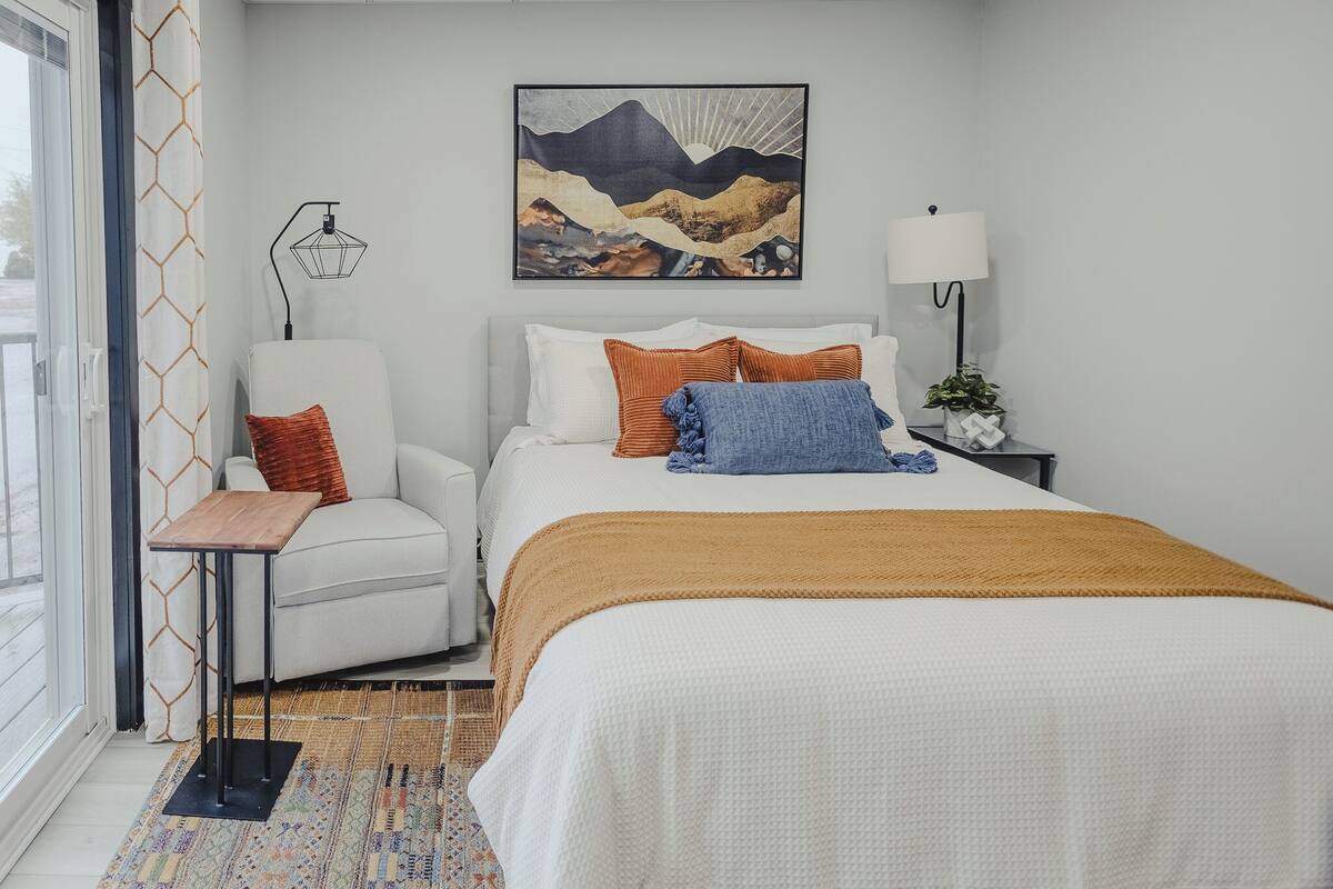 Un dormitorio con cama, una silla, una mesita de noche, lámpara, un cuadro y ventana con cortinas.