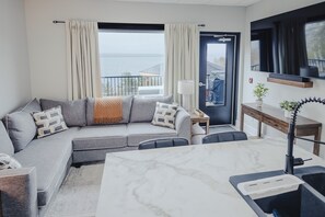 A modern living room with a grey sectional sofa, a marble dining table, a small side table, and a view of the sea through large windows. - Falkenberg Business Centre and Suites (Botwood)