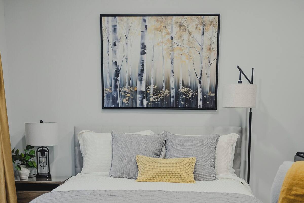Un dormitorio con cama, mesitas de noche, lámpara, silla, alfombra y un cuadro en la pared.